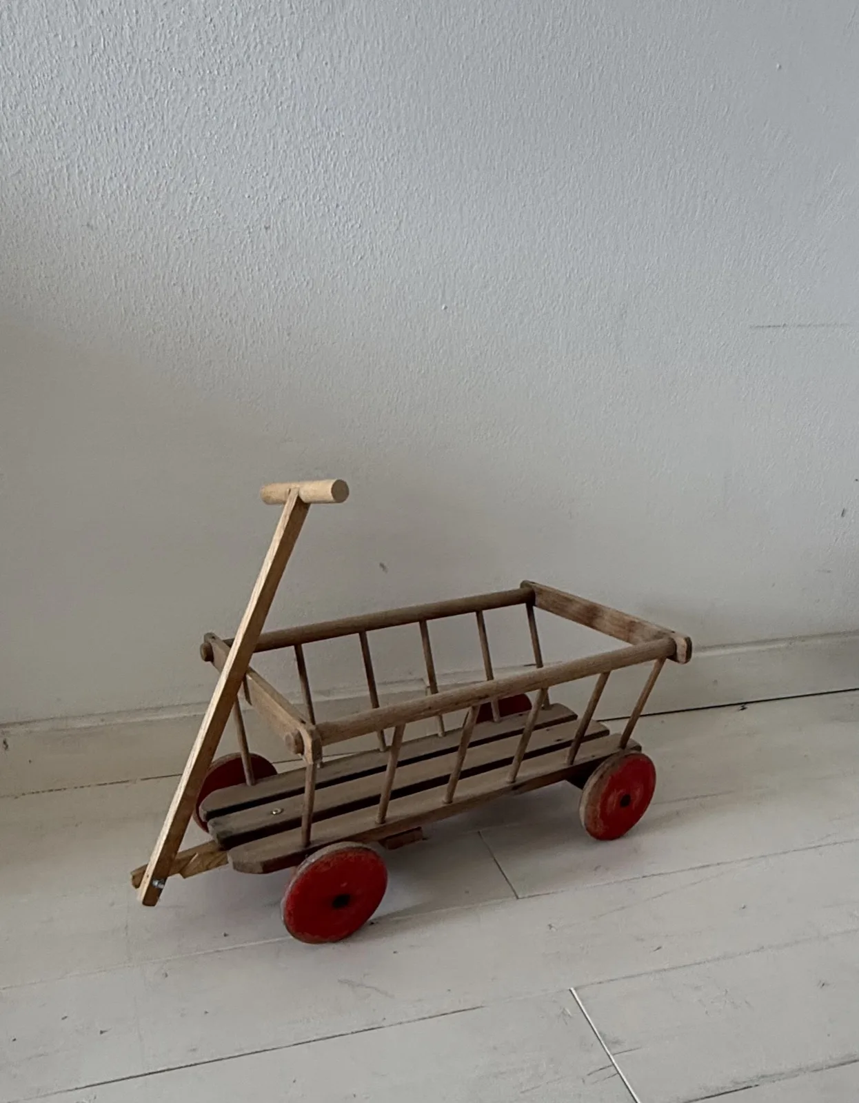 Ein kleiner Leiterwagen aus Holz mit roten Rädern auf hellen Dielen vor einer weißen Wand