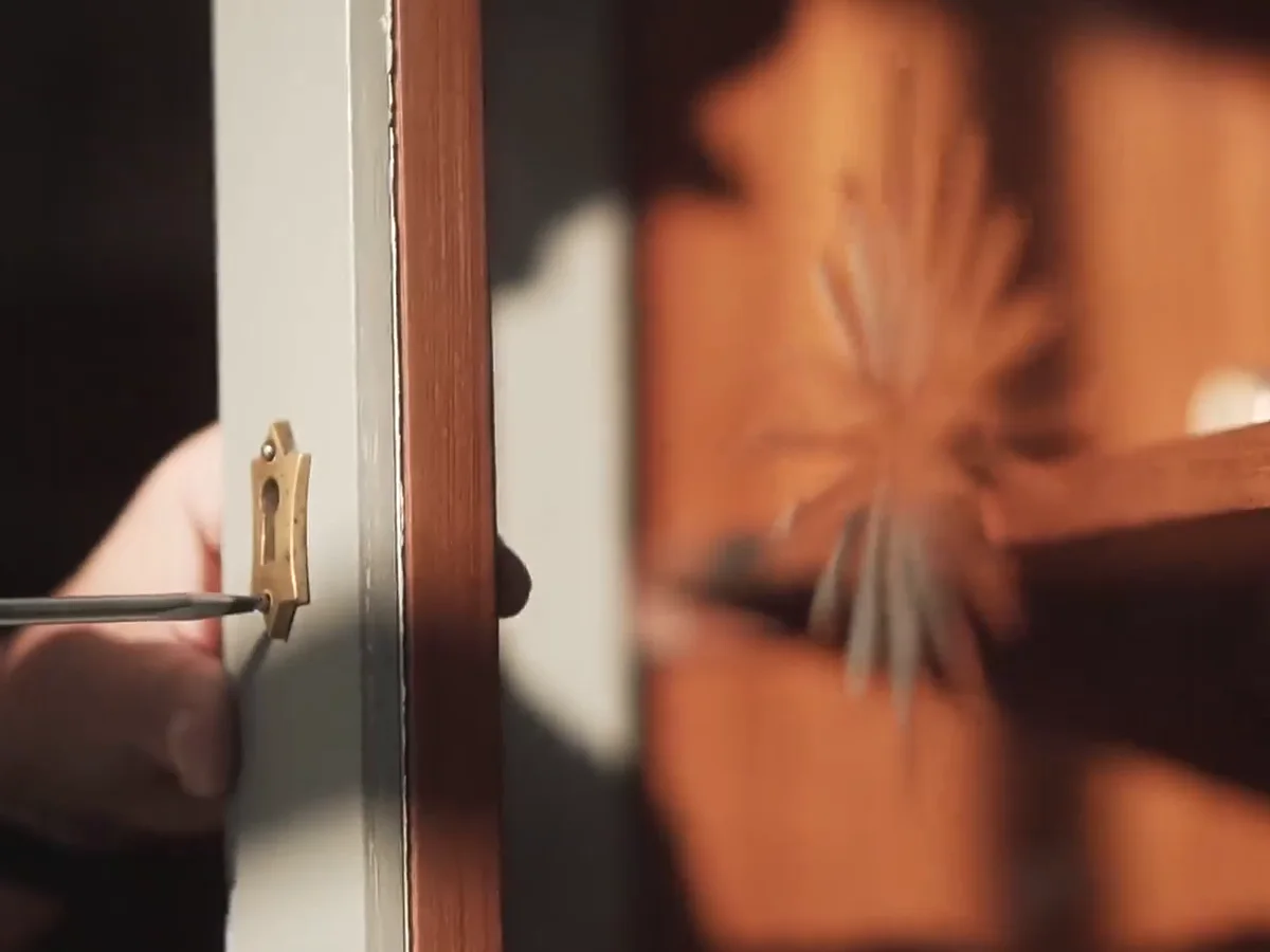 Eine Hand setzt einen Schraubenzieher am Messing-Schlüsselschild einer weißen Kastentür an, dahinter unscharf das Innere des Kastens